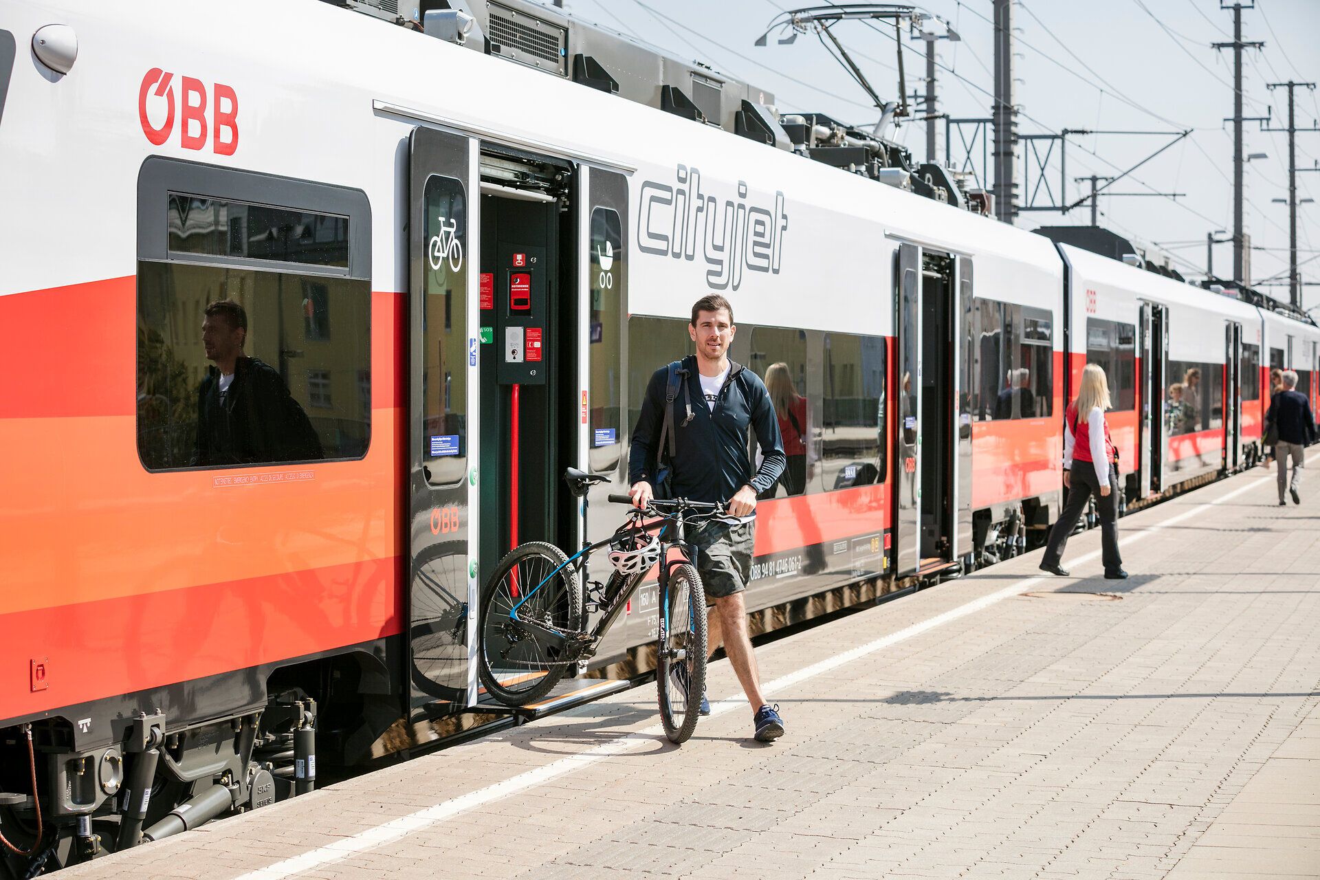 Ein junger Mann steigt mit seinem Fahrrad aus dem modernen Zug, während die Sonne am Himmel strahlt. Die lebendige Atmosphäre des Bahnhofs lädt dazu ein, die Umgebung zu erkunden und die Schönheit der Natur zu genießen.