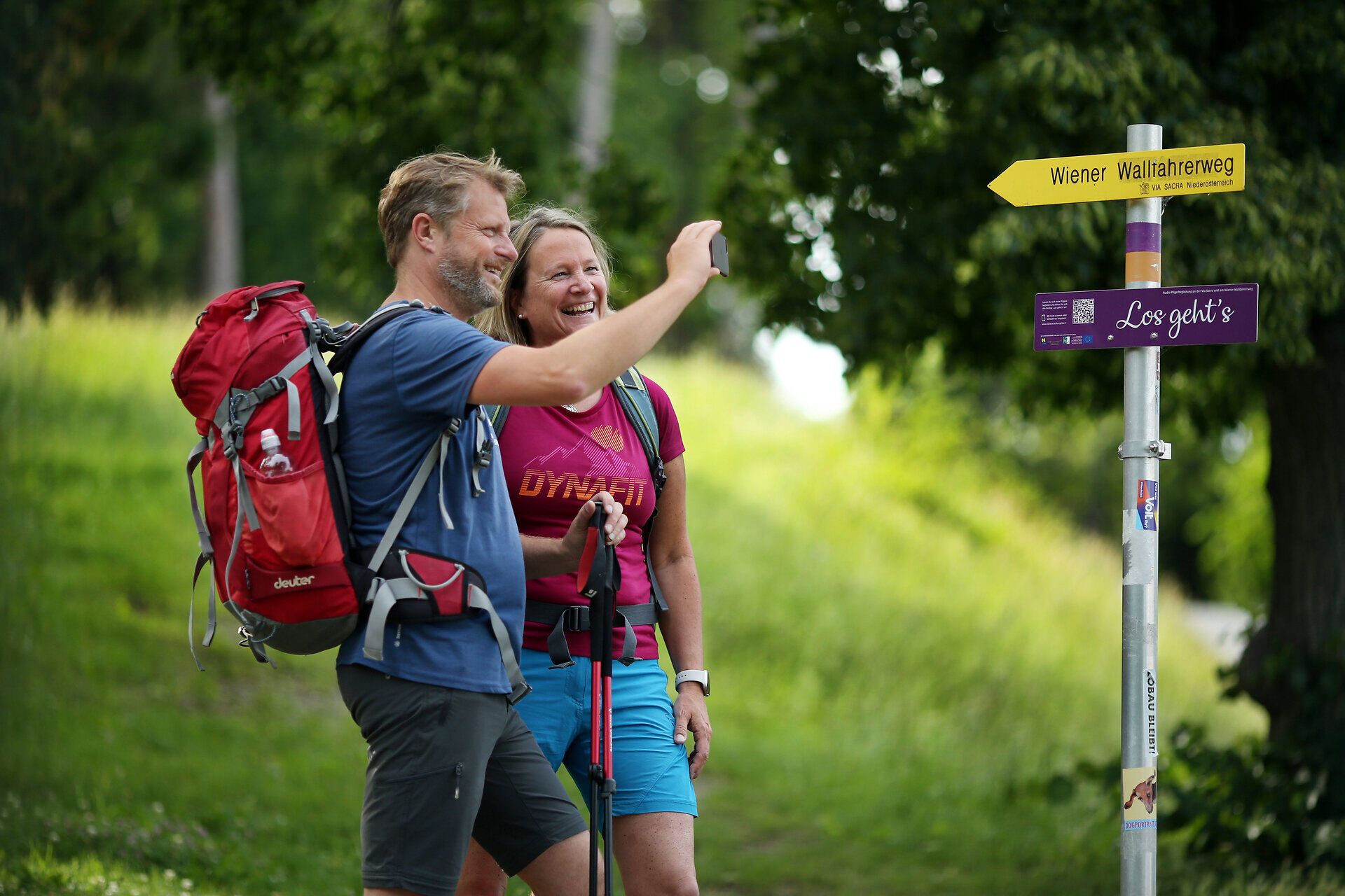 Ein Paar genießt die frische Bergluft und die malerische Landschaft, während sie sich auf dem Wiener Wallfahrerweg befinden. Die strahlende Sonne und die üppigen grünen Wiesen schaffen eine einladende Atmosphäre für Wanderer und Pilger. Hier wird die Schönheit des Sommers in den Bergen spürbar.